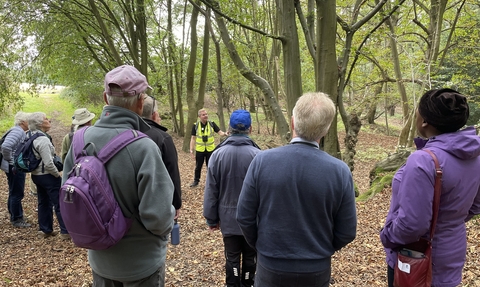 A group of people wearing outdoor clothes gathered in a clearing in a forest in early autumn listening to a man in a reflective jacket who is giving a talk