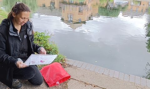 A woman in a raincoat by a river reading instructions from a water testing kit