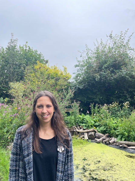 A photograph of Nicola Thompson. She has shoulder length brown hair and is standing in a wildlife garden.