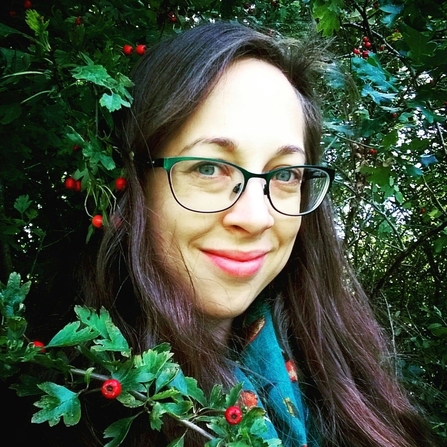 A smiling middle-aged woman with long dark brown hair and glasses is standing in a woodland