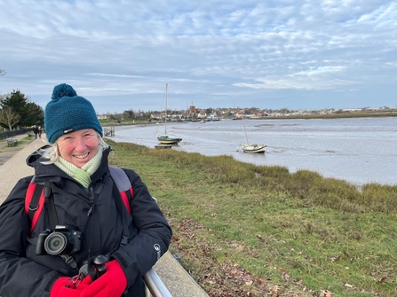 Woman in black coat, green woolly hat and red gloves with a camera round her neck, standing by a grass bank and distant estuary with small boats