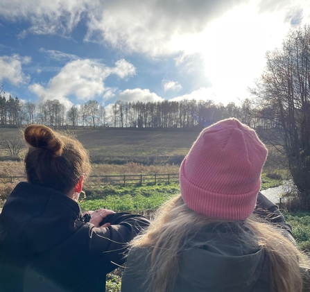 Two people leaning on a fence looking out over grassland to a line of trees on the horizon